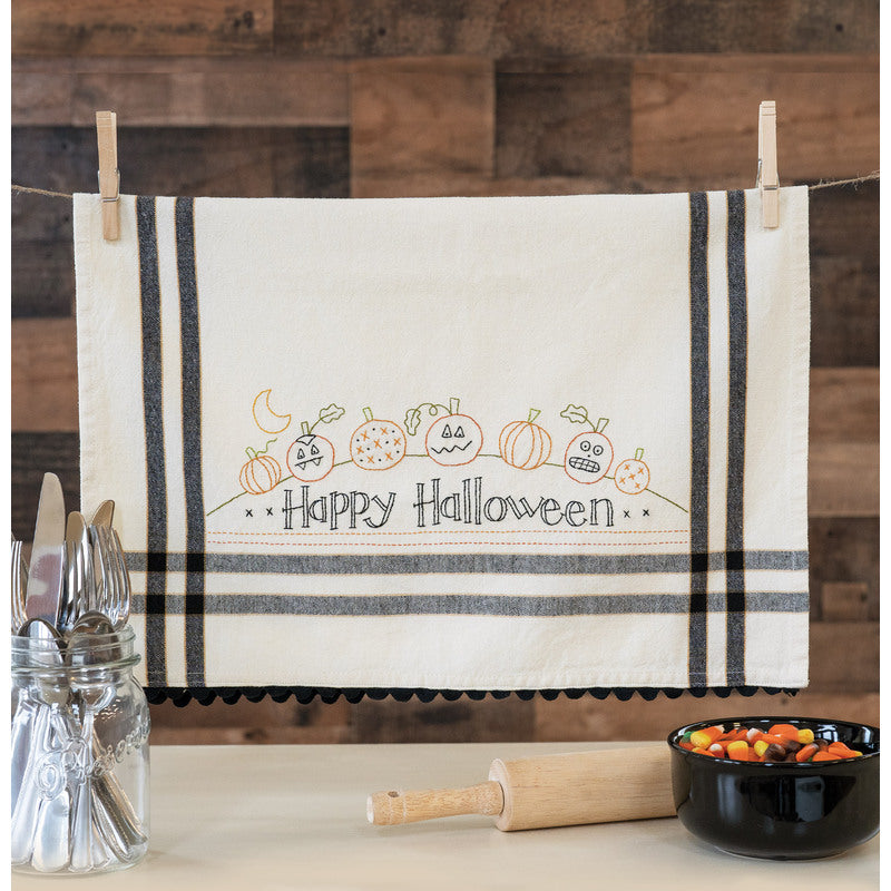 An off-white cotton kitchen towel with black stripes and a 'Happy Halloween' pumpkin embroidery, hanging from a string with wooden clothespins on a rustic wood-paneled wall.