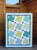 A square quilted blanket or small quilt panel with a geometric star-block pattern in teal, mint green, yellow-green, and white, displayed leaning against a rustic wooden cabin wall.