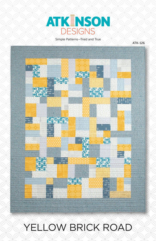 A quilt pattern cover for 'Yellow Brick Road' by ATKINSON DESIGNS, featuring a patchwork quilt with yellow, blue, and gray blocks on a textured gray background.