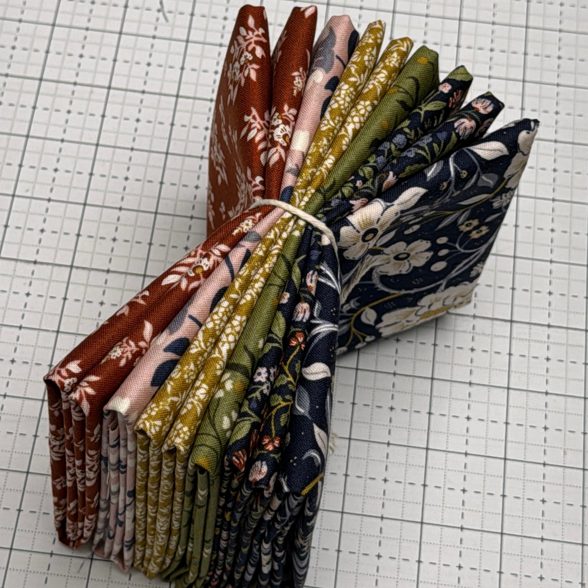A bundle of folded After the Bloom patterned fabric pieces tied with a white string, displayed on a white gridded cutting mat. The fabrics feature various floral and botanical designs in rust red, dusty pink, olive green, mustard yellow, and navy blue.
