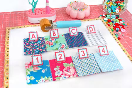 A sewing and fabric organization kit displayed on a white mat, featuring four labeled fabric swatches (A, B, C, D) with different floral and geometric patterns, four numbered fabric swatches (1-4) in blue, pink, yellow, and light blue, a fabric roller, pins, buttons, and other sewing tools on a pink cutting mat background.