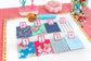 A sewing and fabric organization kit displayed on a white mat, featuring four labeled fabric swatches (A, B, C, D) with different floral and geometric patterns, four numbered fabric swatches (1-4) in blue, pink, yellow, and light blue, a fabric roller, pins, buttons, and other sewing tools on a pink cutting mat background.