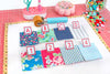 A sewing and fabric organization kit displayed on a white mat, featuring four labeled fabric swatches (A, B, C, D) with different floral and geometric patterns, four numbered fabric swatches (1-4) in blue, pink, yellow, and light blue, a fabric roller, pins, buttons, and other sewing tools on a pink cutting mat background.
