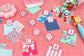 A top-down view of a sewing or quilting project with numbered fabric swatches (1 through 6) labeled with letters A through F on clear blocks, arranged on a pink cutting mat alongside buttons, a pin cushion, and a rotary cutter.