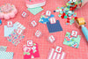 A top-down view of a sewing or quilting project with numbered fabric swatches (1 through 6) labeled with letters A through F on clear blocks, arranged on a pink cutting mat alongside buttons, a pin cushion, and a rotary cutter.