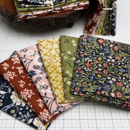 A set of folded fabric swatches  from the After the Bloom collection with various floral and botanical patterns in earthy tones, arranged on a white gridded cutting mat with more stacks in the background.