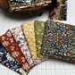 A set of folded fabric swatches  from the After the Bloom collection with various floral and botanical patterns in earthy tones, arranged on a white gridded cutting mat with more stacks in the background.