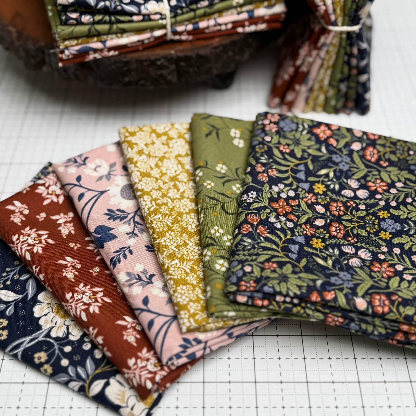 A set of folded fabric swatches  from the After the Bloom collection with various floral and botanical patterns in earthy tones, arranged on a white gridded cutting mat with more stacks in the background.