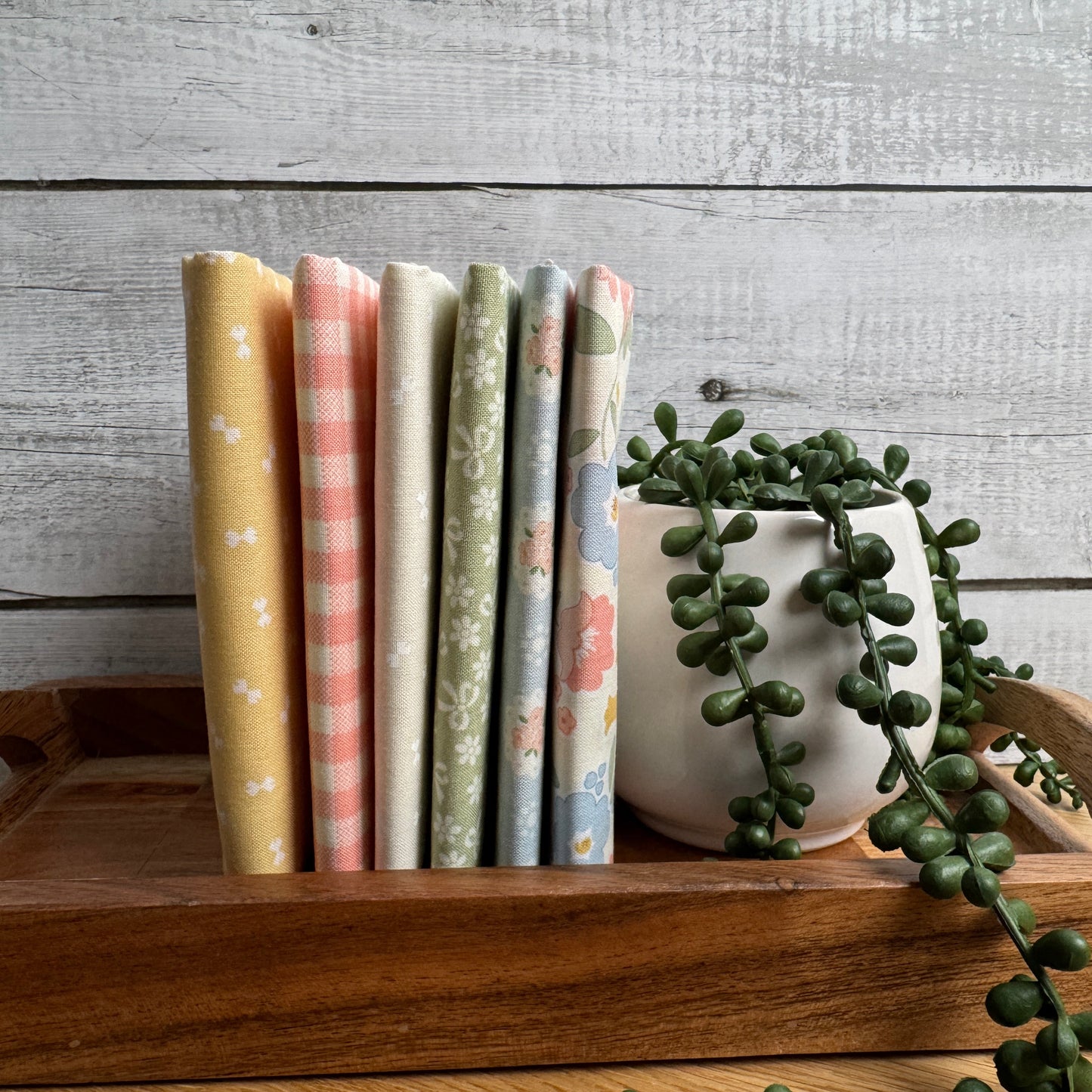 Six folded fat quarters with pastel patterns (check, gingham, floral, and stripes) are arranged in a wooden tray next to a potted trailing green plant against a rustic gray wood plank background.