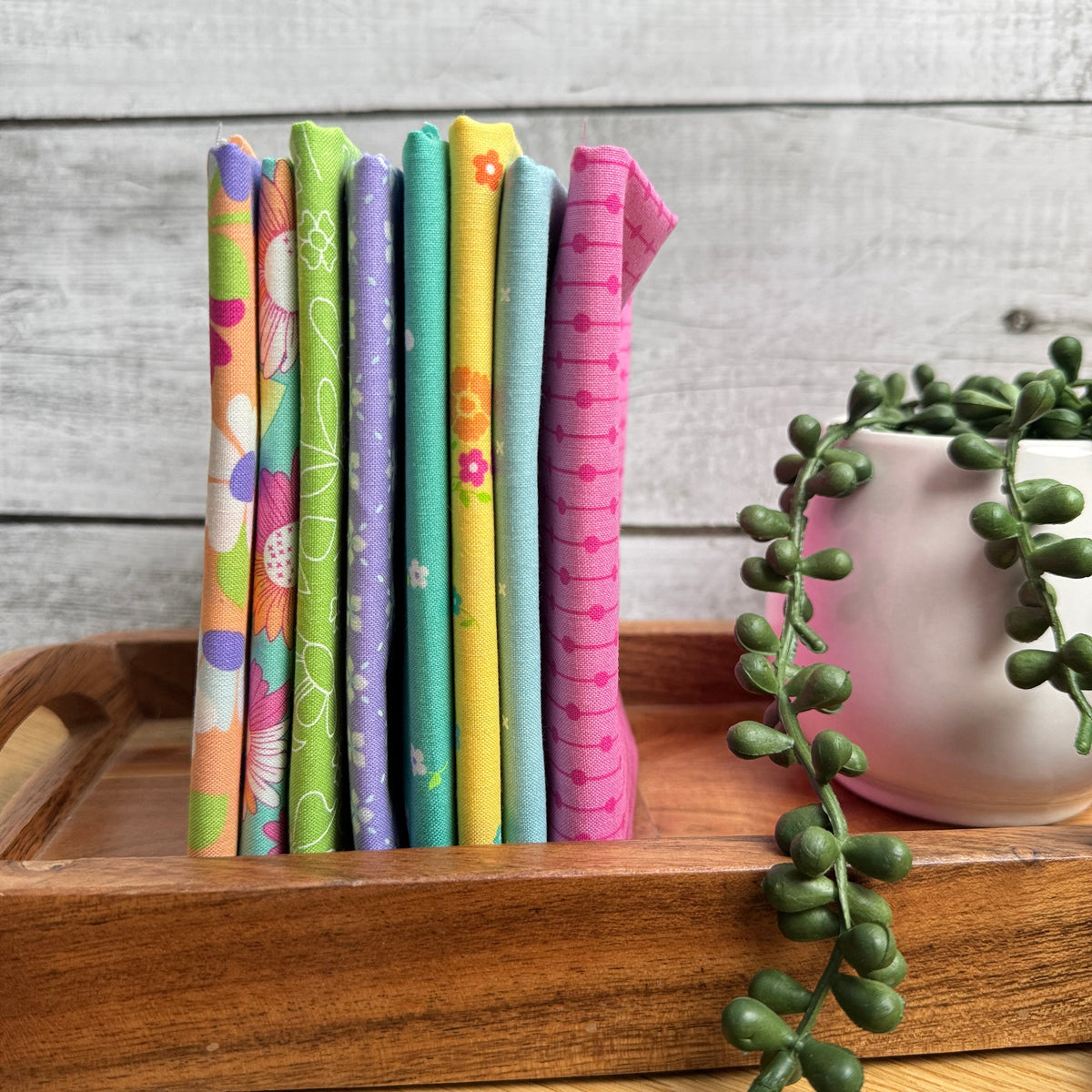 Joysome Cantaloupe 8-piece fat quarter bundle in orange tones, displayed standing on edge on a decorative tray beside a faux plant.