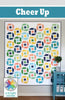 A quilted wall hanging with a repeating geometric patchwork pattern in multiple colors, shown in a styled room scene with a teal cabinet and a plant. The image includes a teal banner at the top reading 'Cheer Up' and a 'a bright corner' logo in the lower left.