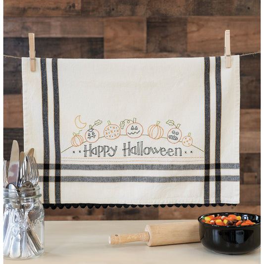 An off-white cotton kitchen towel with black stripes and a 'Happy Halloween' pumpkin embroidery, hanging from a string with wooden clothespins on a rustic wood-paneled wall.