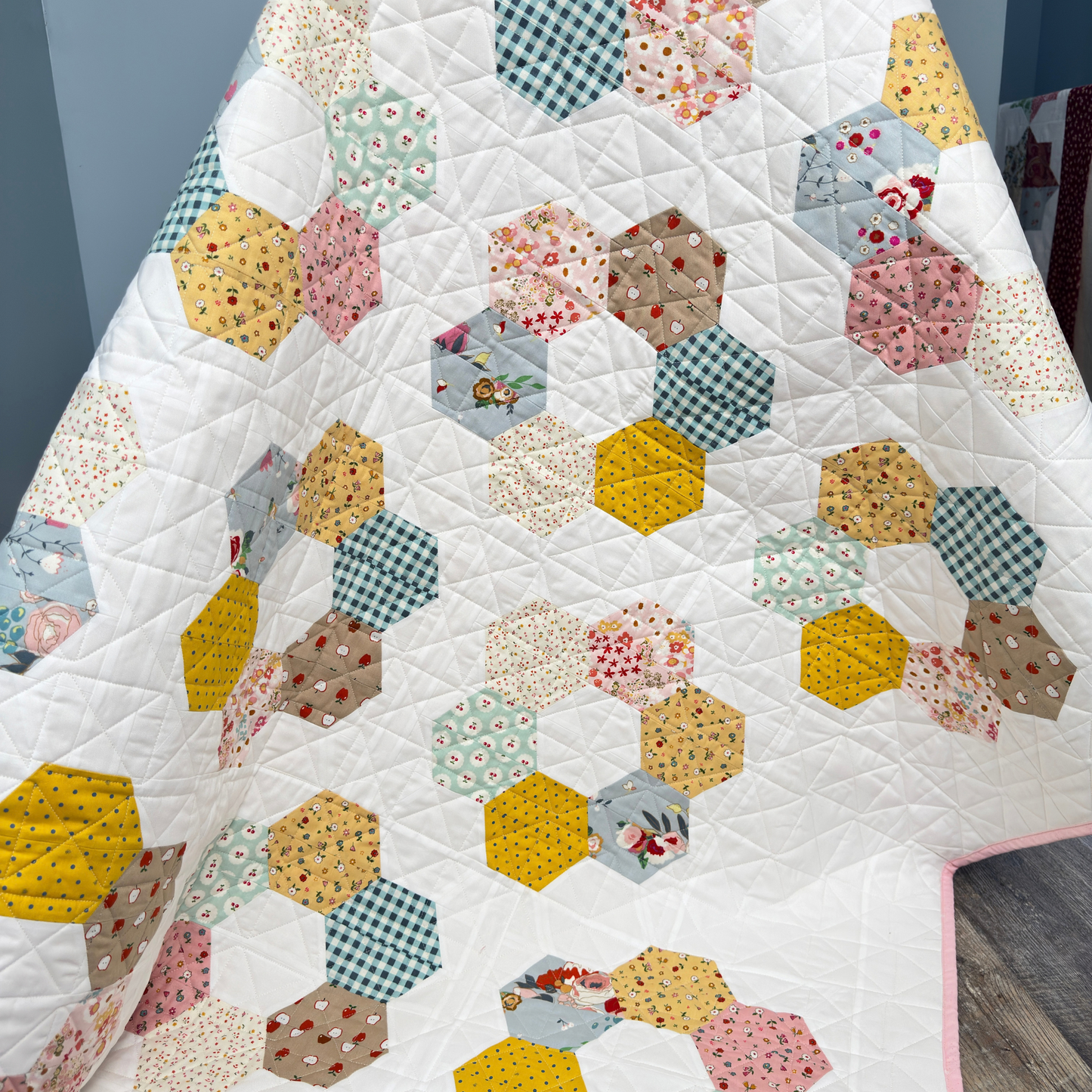 A handmade hexagon patchwork quilt draped over a chair, featuring a white background with various colorful fabric hexagons in yellow, pink, blue, green, and brown patterns from the Cotton Nook fabric collection by Minki Kim. 