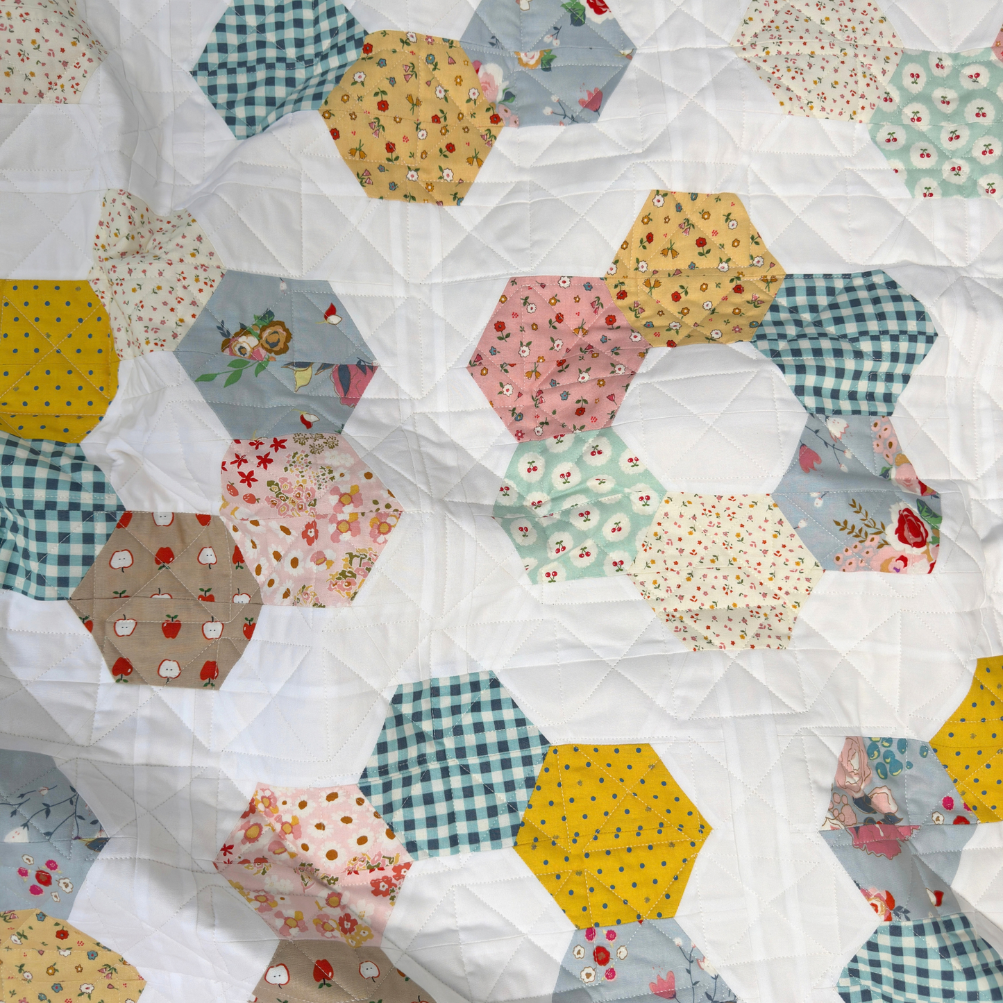 A close-up of a handmade quilt featuring hexagonal patches made from various patterned fabrics, including gingham, floral prints, and solid colors, stitched together on a white background.