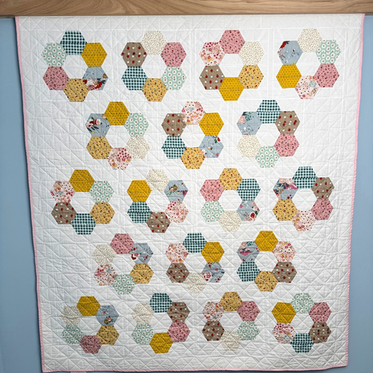 A handmade quilted patchwork quilt made with Cotton Nook fabric featuring repeating rows of hexagonal rings made from various patterned fabric pieces in yellow, pink, blue, green, and brown, displayed against a light blue wall.