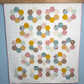 A handmade quilted patchwork quilt made with Cotton Nook fabric featuring repeating rows of hexagonal rings made from various patterned fabric pieces in yellow, pink, blue, green, and brown, displayed against a light blue wall.
