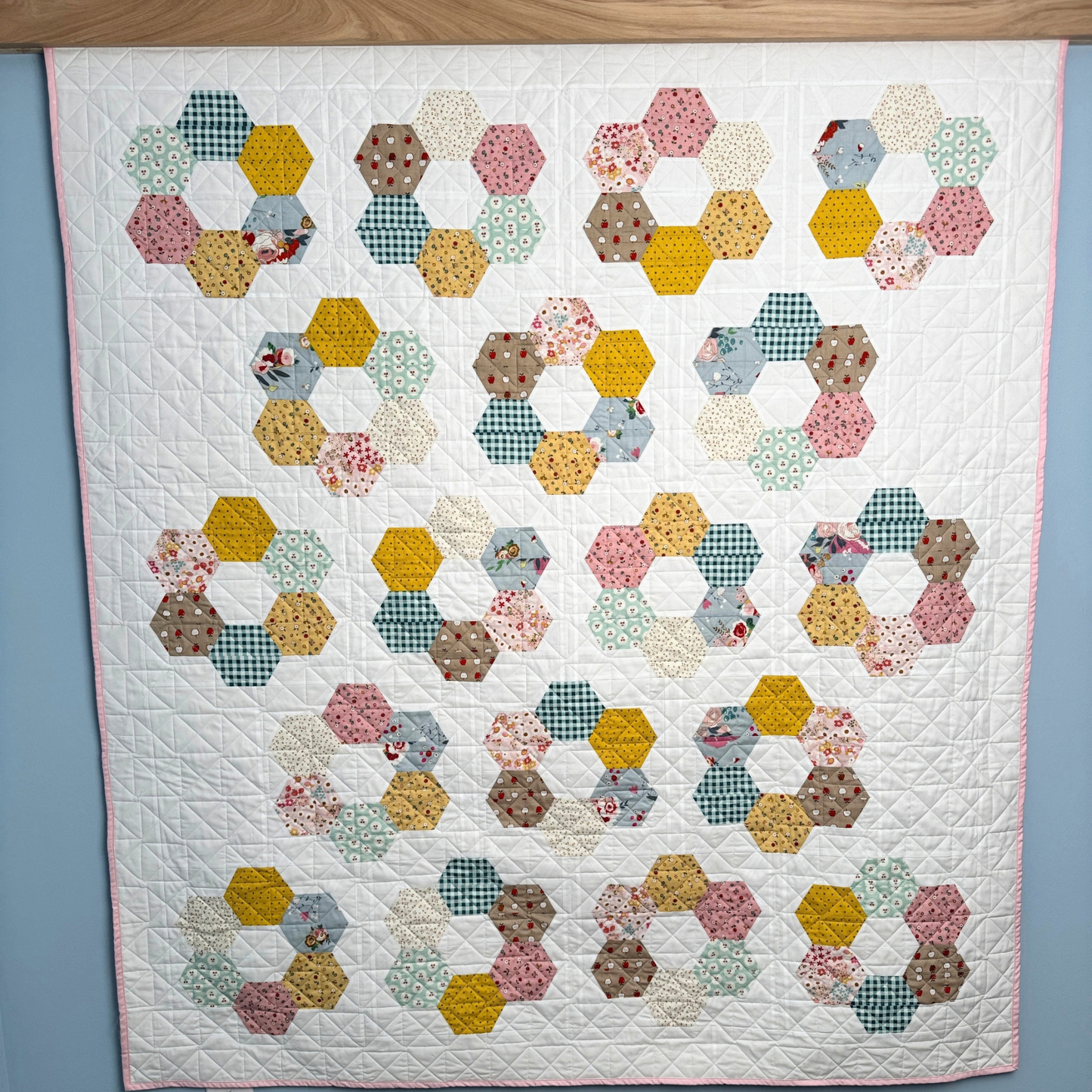 A handmade quilted patchwork quilt made with Cotton Nook fabric featuring repeating rows of hexagonal rings made from various patterned fabric pieces in yellow, pink, blue, green, and brown, displayed against a light blue wall.