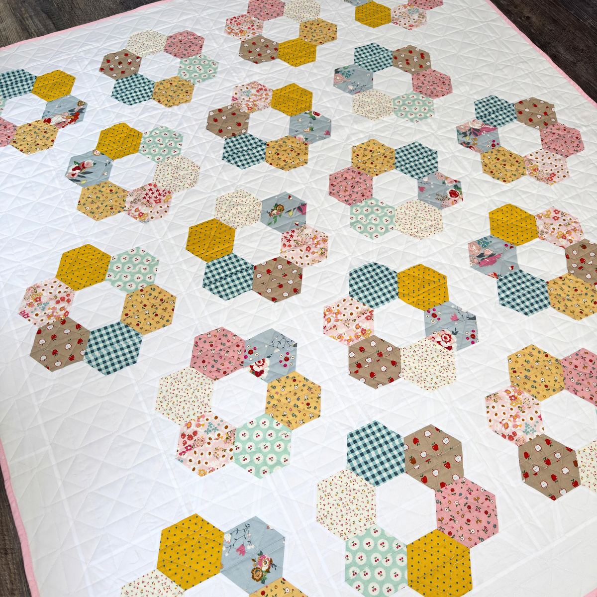 A handmade hexagonal patchwork quilt laid flat on a dark wood floor. The quilt features a white background with rows of colorful fabric hexagons in yellow, green, pink, blue, and brown, including gingham, floral, and polka dot patterns.