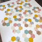 A handmade hexagonal patchwork quilt laid flat on a dark wood floor. The quilt features a white background with rows of colorful fabric hexagons in yellow, green, pink, blue, and brown, including gingham, floral, and polka dot patterns.
