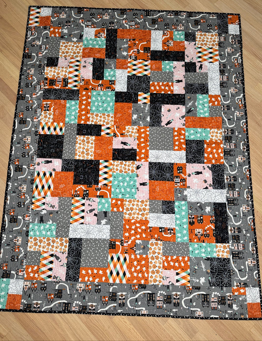 A rectangular patchwork quilt laid flat on a light wood floor. The quilt features a patchwork design with orange, black, gray, white, and mint green blocks, with a dark gray border printed with small cartoon characters.