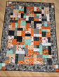 A rectangular patchwork quilt laid flat on a light wood floor. The quilt features a patchwork design with orange, black, gray, white, and mint green blocks, with a dark gray border printed with small cartoon characters.