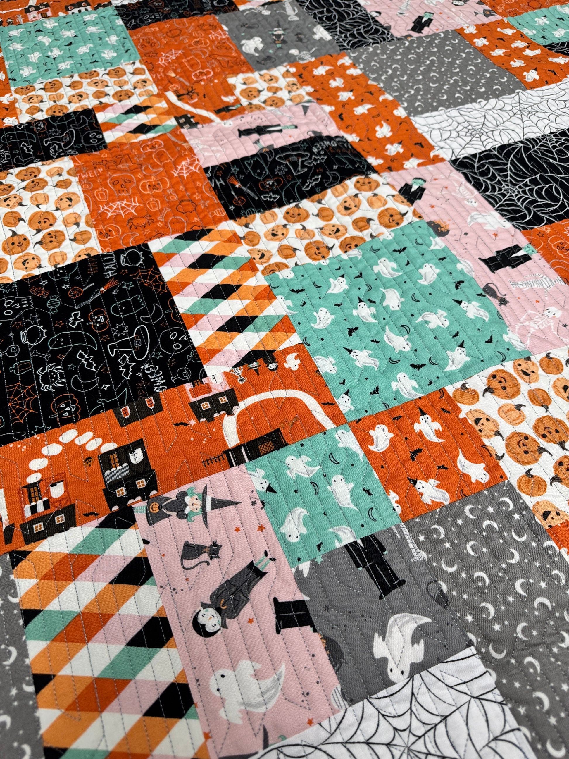 A close-up of a patchwork quilt featuring various Halloween-themed fabric panels with orange, pink, teal, black, and gray patterns of pumpkins, ghosts, witches, and spiderwebs.