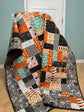 A patchwork quilt draped over a white chair in a room with a light blue wall and wood floor. The quilt features a mix of orange, gray, black, pink, and mint green patterned squares, including a bus route motif on the border.