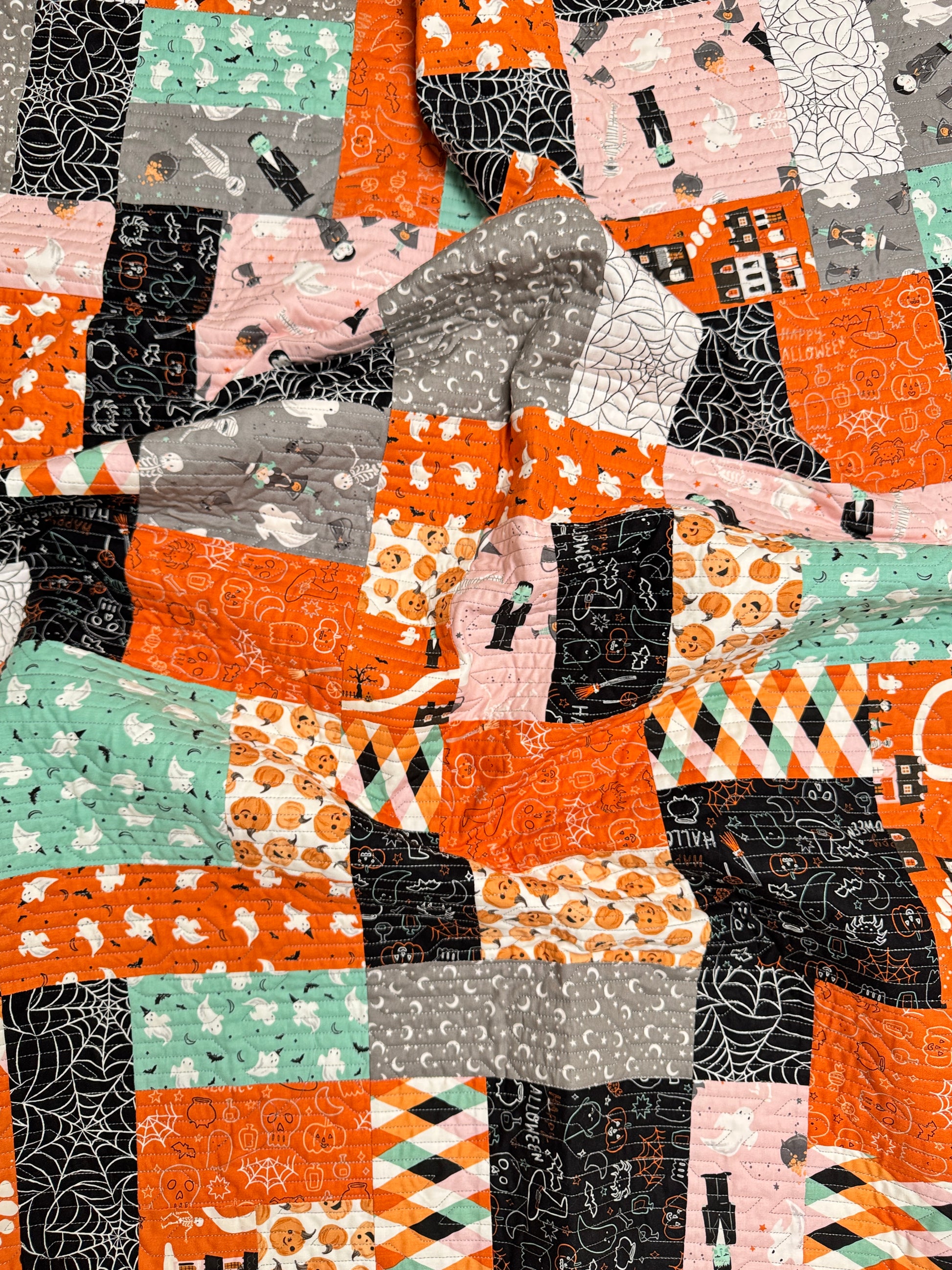 A close-up of a patchwork quilt featuring Halloween-themed fabrics with orange, black, pink, gray, and mint green patterns of pumpkins, ghosts, spiderwebs, and haunted houses.