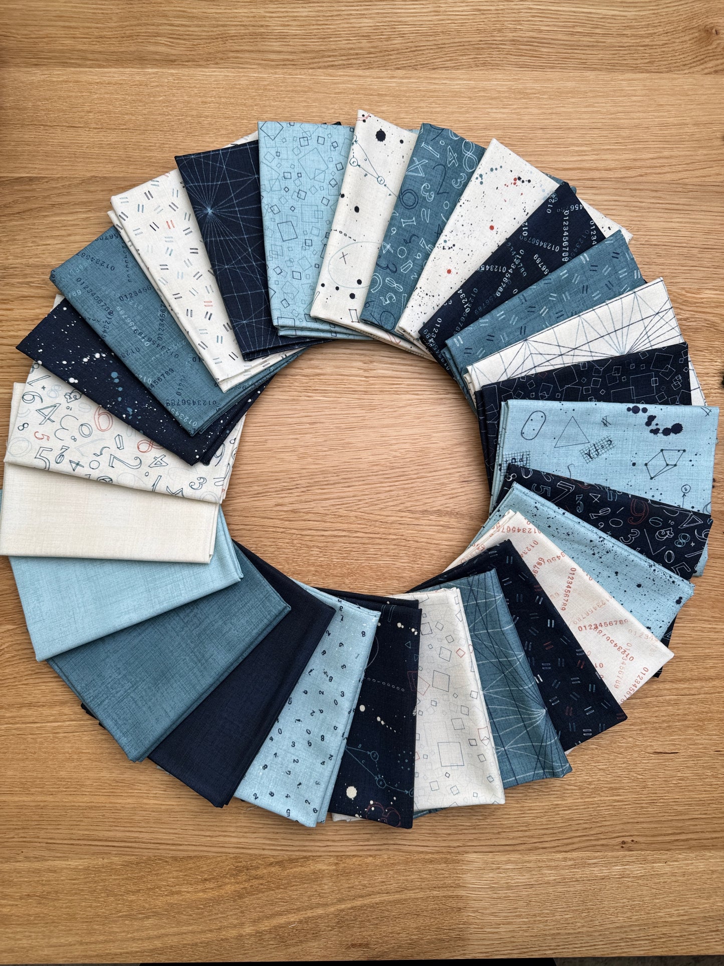 A circular arrangement of Data by Janet Clare fabric swatches in various shades of blue, navy, cream, and white with celestial and geometric patterns, displayed on a light brown wooden surface.