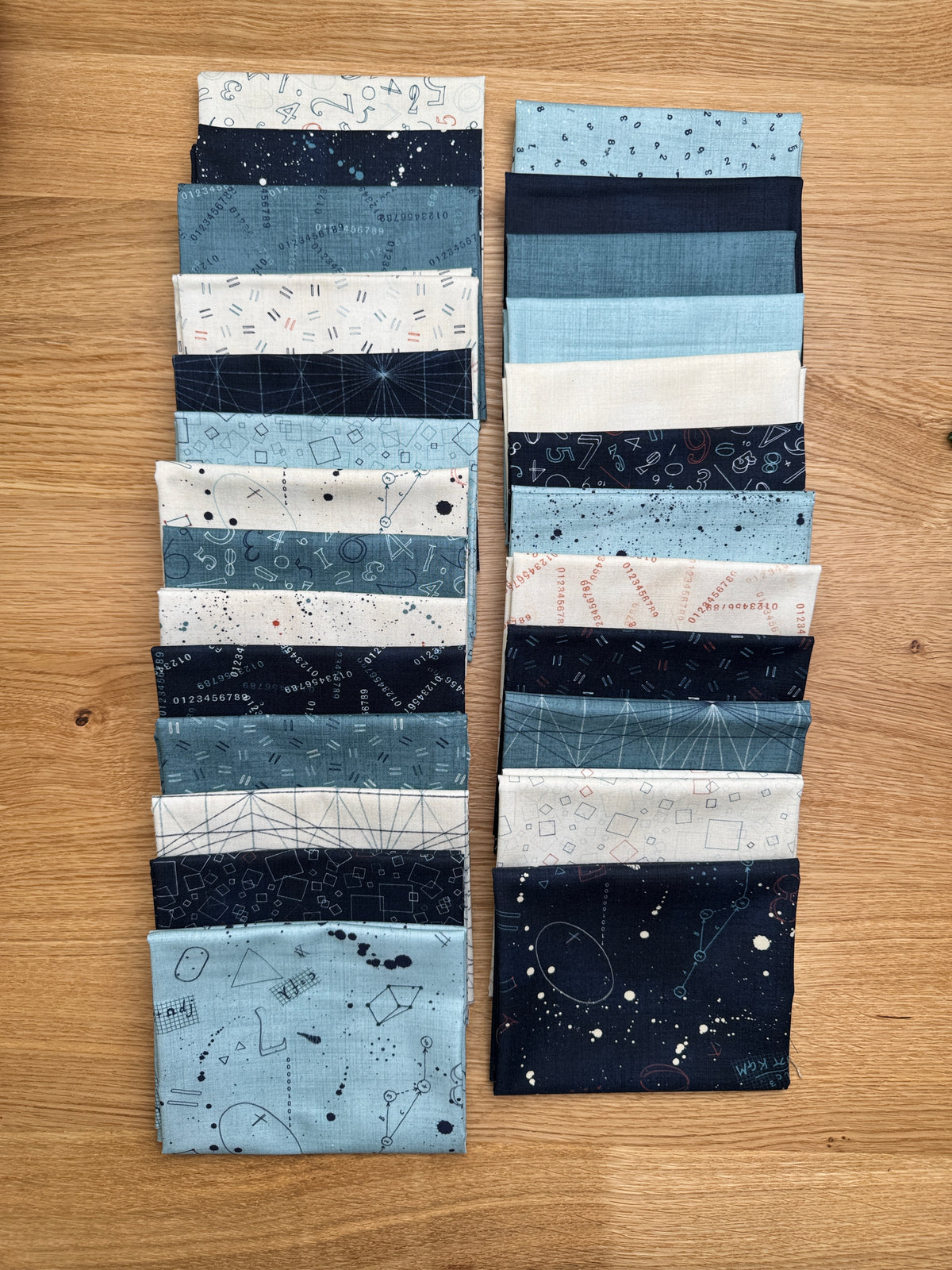 Two vertical stacks of Data fabric swatches by Janet Clare in shades of blue, navy, white, and gray with various printed patterns, arranged on a wooden surface.