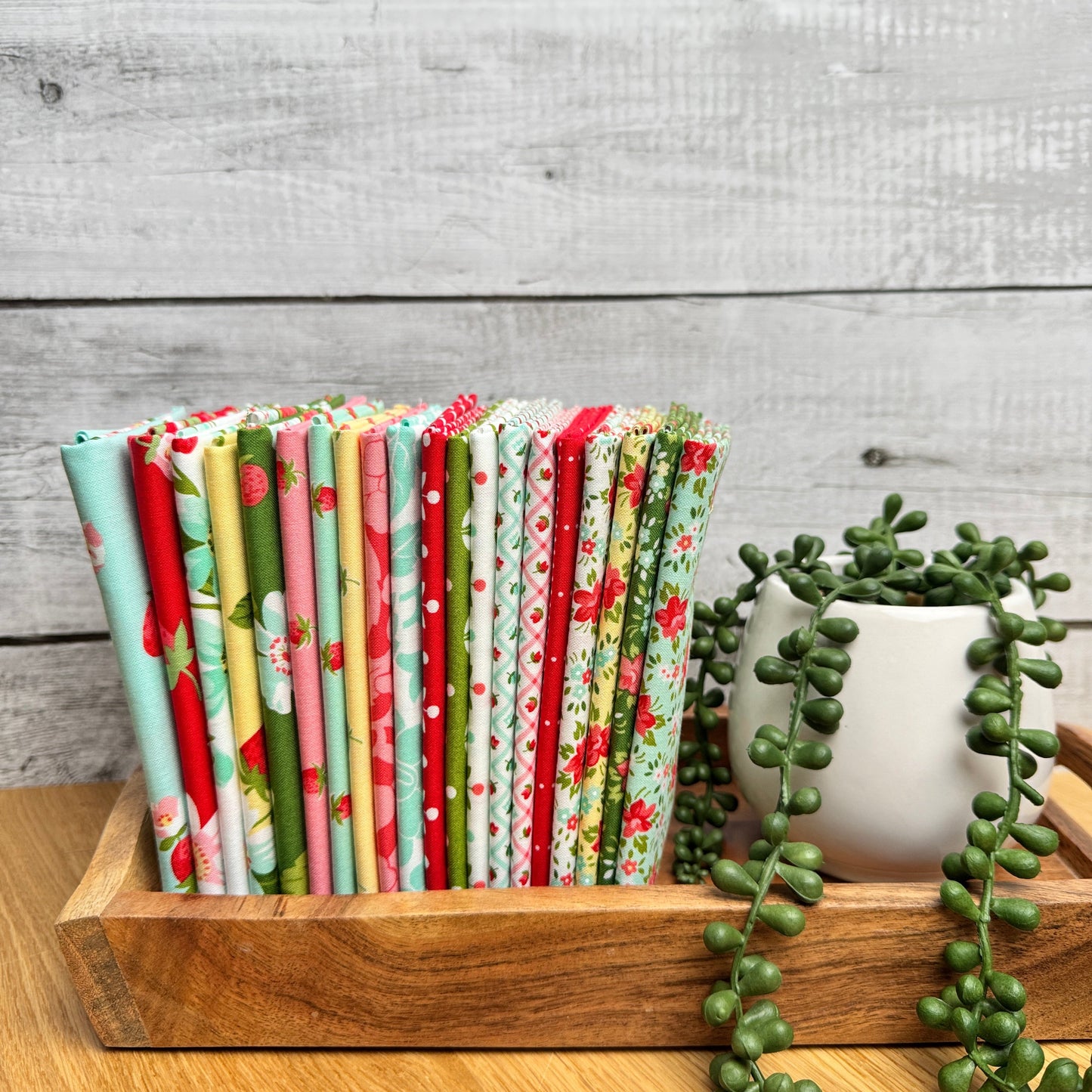 20 piece fat quarters from the Bloomsberry collection by Camille Roskelley lined up on a wooden tray next to a fake plant.  