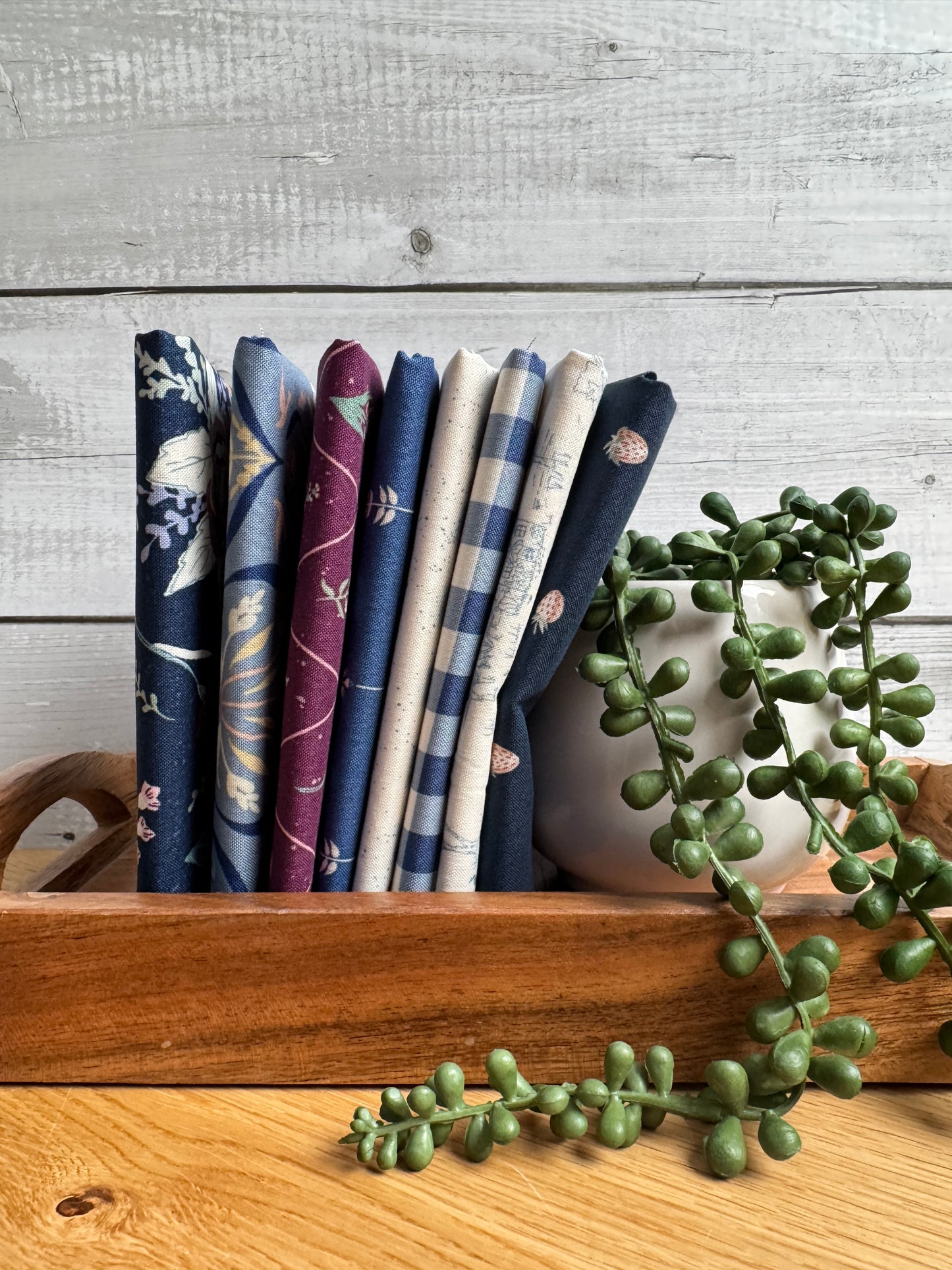 Blue Hedgerow Haven petite fat quarter bundle standing on end on a wooden tray next to a plant
