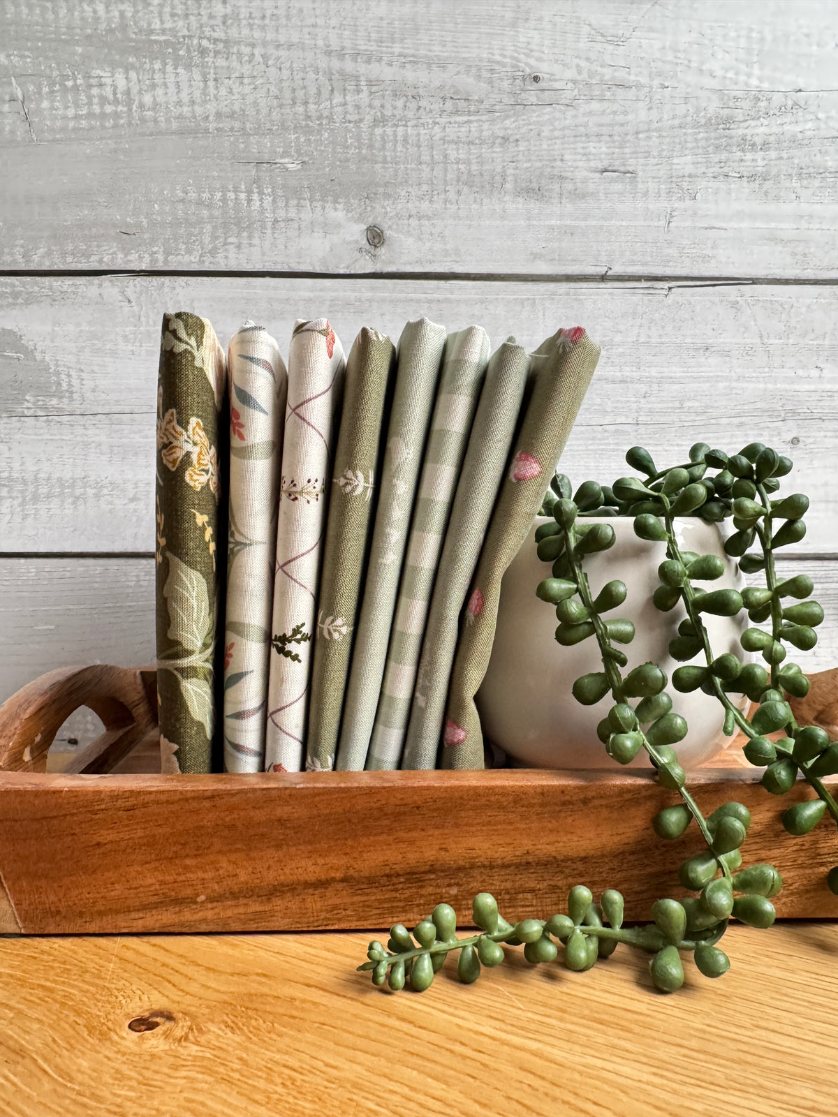 Green Hedgerow Haven petite fat quarter bundle standing on end on a wooden tray next to a plant