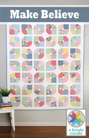 Make Believe quilt pattern cover.  A quilted wall hanging or patchwork textile displayed indoors, featuring a grid of colorful geometric and floral fabric squares on a white base. The top has a blue-gray banner with the text 'Make Believe,' and a 'a bright corner' logo appears in the lower right corner.
