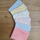 Six folded fat quarters  in pastel colors and patterns, arranged in a fan shape on a light brown wooden surface. The fabrics feature floral, polka dot, and striped designs in pink, blue, yellow, and white.