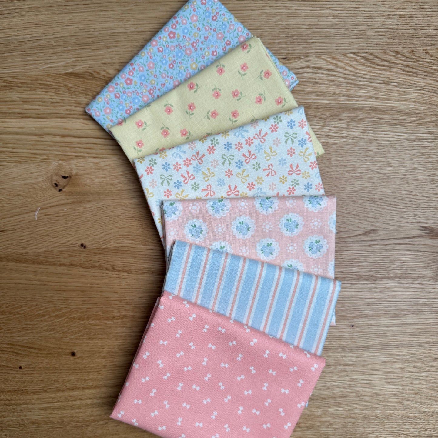Six folded fat quarters  in pastel colors and patterns, arranged in a fan shape on a light brown wooden surface. The fabrics feature floral, polka dot, and striped designs in pink, blue, yellow, and white.