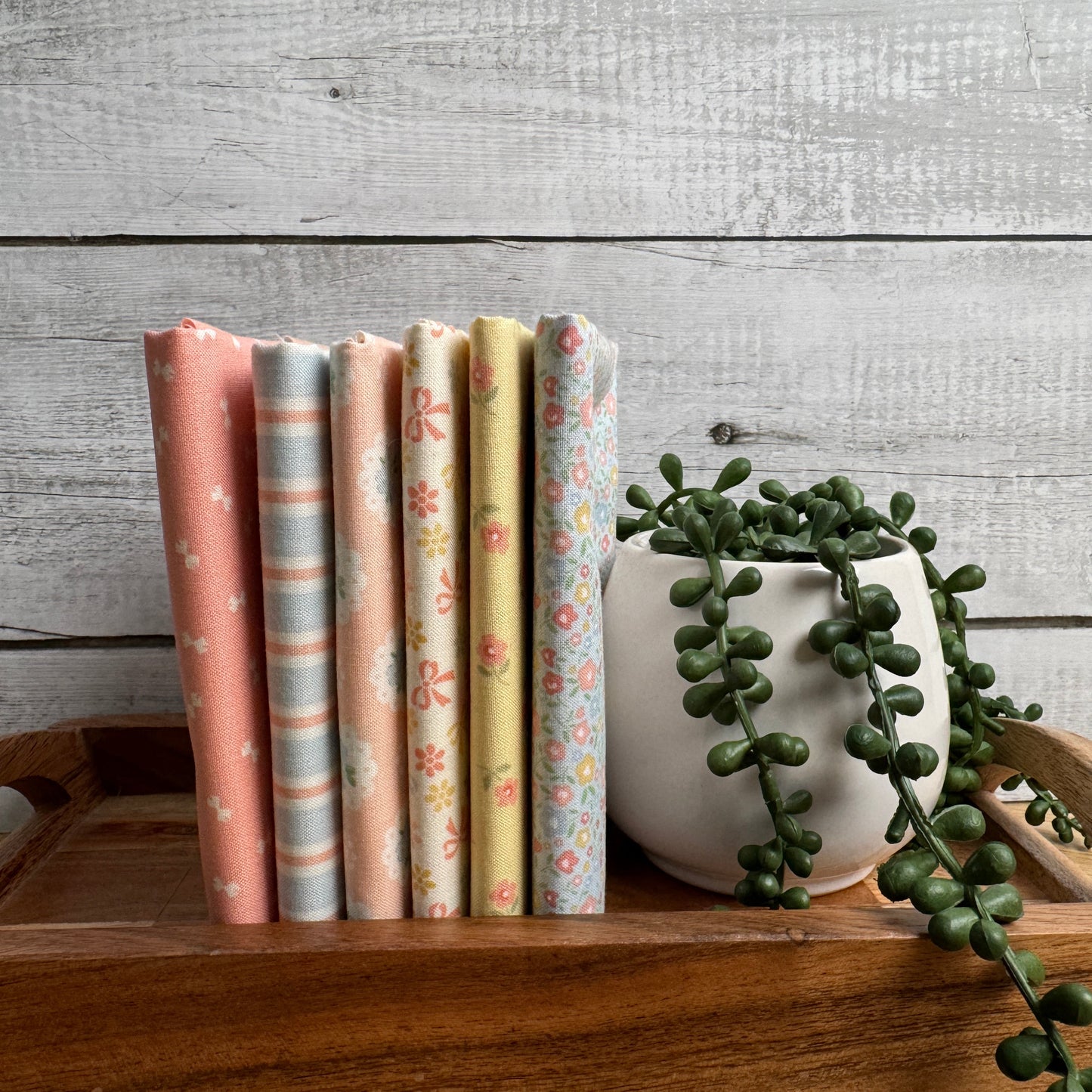 Six folded fabric samples with pastel patterns (stripes, florals, polka dots) are displayed on a wooden tray next to a white ceramic pot with a trailing green plant, set against a rustic gray wood plank background.