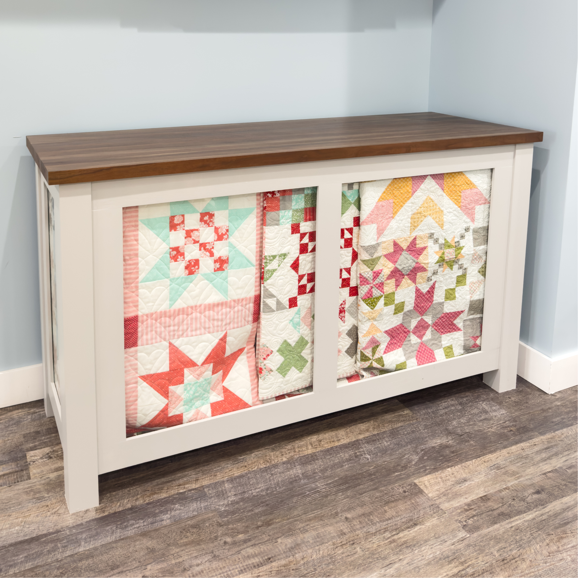 A white storage cabinet with a dark brown wood top, featuring two glass-front doors displaying colorful quilted fabrics, set against a light blue wall and wood-look floor.