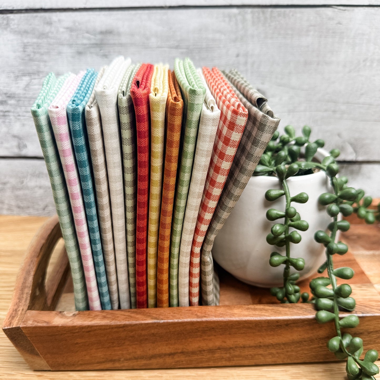 Thirteen Quiltessential Minis check fat quarters standing on end on a tray beside an artificial plant.