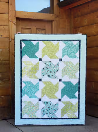 A square quilted blanket or small quilt panel with a geometric star-block pattern in teal, mint green, yellow-green, and white, displayed leaning against a rustic wooden cabin wall.