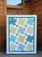 A square quilted blanket or small quilt panel with a geometric star-block pattern in teal, mint green, yellow-green, and white, displayed leaning against a rustic wooden cabin wall.